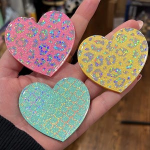 May include: Three heart-shaped resin ornaments with iridescent glitter. The hearts are pink, yellow, and teal with a leopard print design.