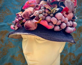May include: A decorative hat with a dark blue brim and a beige underside. The hat is adorned with clusters of purple, pink, and red fabric flowers and grape-like embellishments, creating a whimsical and artistic design.