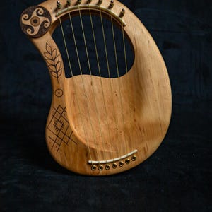 Könnte beinhalten: Eine hellbraune Holzbogenharfe mit goldenen Saiten und dekorativen Schnitzereien. Das Instrument hat eine geschwungene Form mit einem kreisförmigen Design oben und geometrischen Mustern auf dem Korpus. Ca. 50 cm lang.