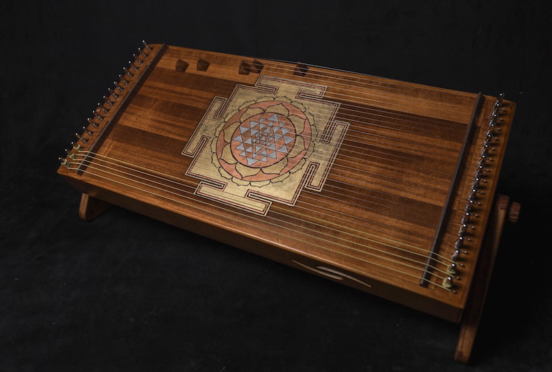May include: A wooden stringed instrument with a decorative geometric design in gold, pink, and blue. The instrument is set on a wooden stand, displaying its warm wood tones. The instrument is likely a type of zither.