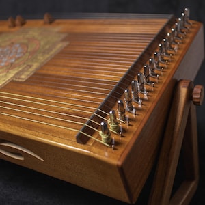 May include: A wooden stringed instrument, possibly a zither, with a decorative pattern on its surface. The instrument is supported by a wooden stand. Metal tuning pegs are visible. The wood has a warm, brown tone.