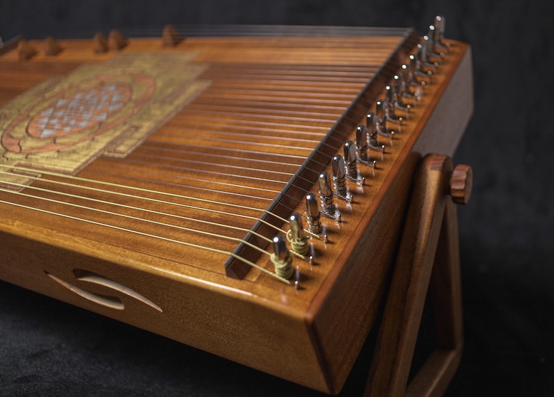May include: A wooden stringed instrument, possibly a zither, with a decorative pattern on its surface. The instrument is supported by a wooden stand. Metal tuning pegs are visible. The wood has a warm, brown tone.