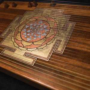 May include: A wooden stringed instrument with a decorative Sri Yantra design. The instrument features gold, copper, and silver accents. The wood grain is visible, and strings run across the surface.