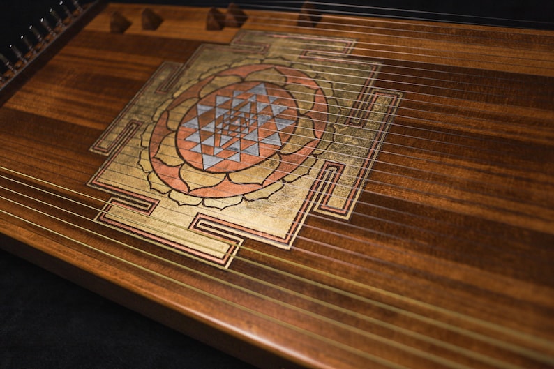 May include: A wooden stringed instrument with a decorative Sri Yantra design. The instrument features gold, copper, and silver accents. The wood grain is visible, and strings run across the surface.