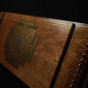 May include: A wooden stringed instrument with a decorative geometric design in the center. The instrument has many strings and tuning pegs along the sides. The wood has a warm, natural tone, and the design features shades of orange, blue, and gold.