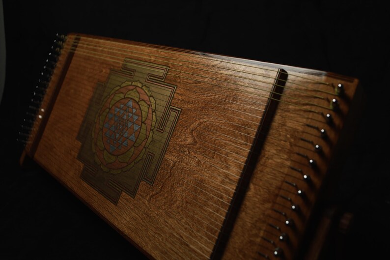May include: A wooden stringed instrument with a decorative geometric design in the center. The instrument has many strings and tuning pegs along the sides. The wood has a warm, natural tone, and the design features shades of orange, blue, and gold.