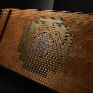 May include: A wooden stringed instrument, possibly a zither, with a decorative central design in gold, pink, and silver. The instrument has a warm brown finish and numerous strings stretched across its surface. The design is geometric.