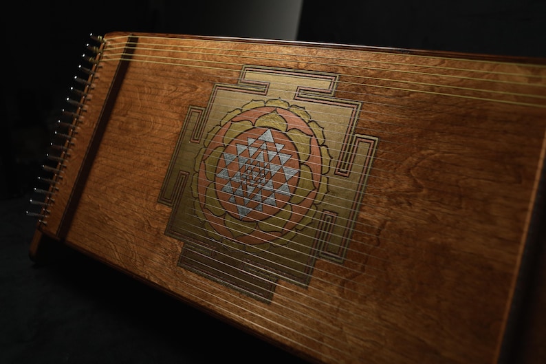 May include: A wooden stringed instrument, possibly a zither, with a decorative central design in gold, pink, and silver. The instrument has a warm brown finish and numerous strings stretched across its surface. The design is geometric.