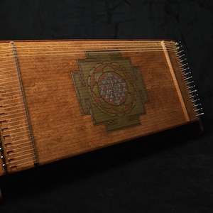 May include: A wooden hammered dulcimer with a central geometric design. The instrument features numerous strings and a wooden stand. The dulcimer is a warm brown colour, set against a dark background. The instrument is approximately 76 cm long.