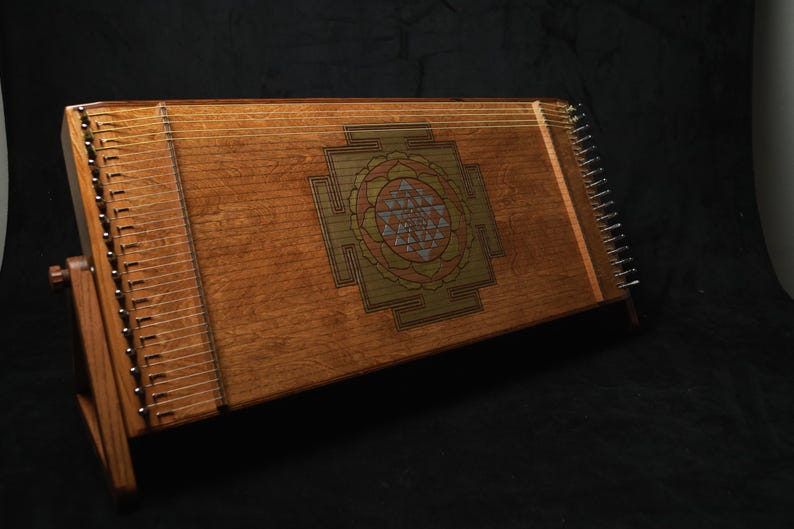 May include: A wooden hammered dulcimer with a central geometric design. The instrument features numerous strings and a wooden stand. The dulcimer is a warm brown colour, set against a dark background. The instrument is approximately 76 cm long.