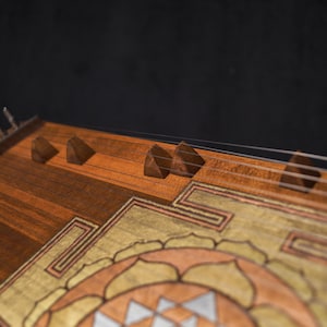 May include: A close-up shot of a wooden hammered dulcimer. The instrument has a detailed design with brown, gold, and silver tones. The strings are visible, stretched across the soundboard, and the instrument is set against a dark background.