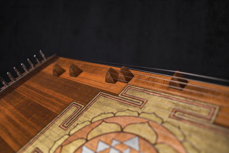 May include: A close-up shot of a wooden hammered dulcimer. The instrument has a detailed design with brown, gold, and silver tones. The strings are visible, stretched across the soundboard, and the instrument is set against a dark background.