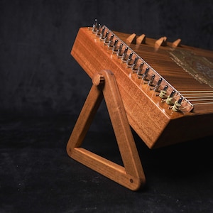 May include: A wooden hammered dulcimer, a stringed musical instrument, is displayed on a wooden stand. The instrument has a rich, brown finish with visible strings and tuning pegs. The stand is a simple, triangular design.