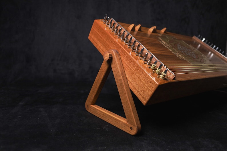 May include: A wooden hammered dulcimer, a stringed musical instrument, is displayed on a wooden stand. The instrument has a rich, brown finish with visible strings and tuning pegs. The stand is a simple, triangular design.
