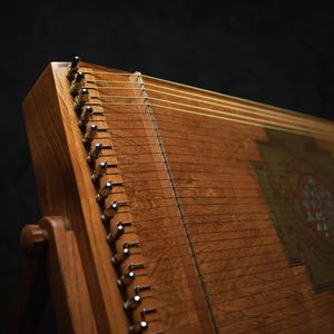May include: A wooden stringed instrument, possibly a zither, in shades of brown. The strings are stretched across the wooden soundboard, with metal tuning pegs. The instrument is set against a dark background.
