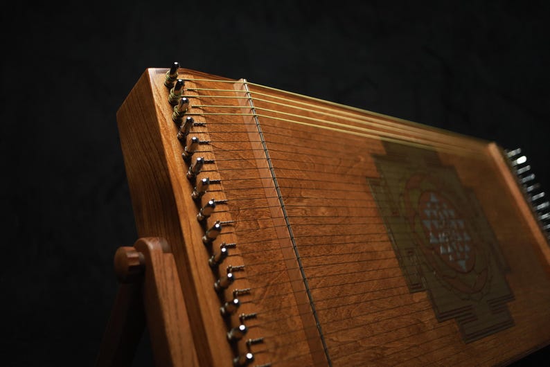 May include: A wooden stringed instrument, possibly a zither, in shades of brown. The strings are stretched across the wooden soundboard, with metal tuning pegs. The instrument is set against a dark background.