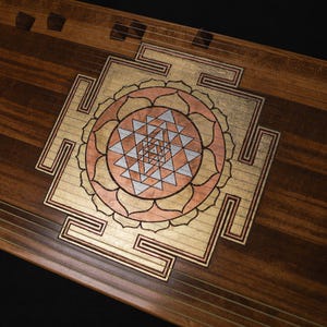 May include: A wooden stringed instrument with an inlaid design. The design features a central geometric pattern in gold, copper, and silver tones, surrounded by a floral-like motif. The instrument's wood grain is visible.