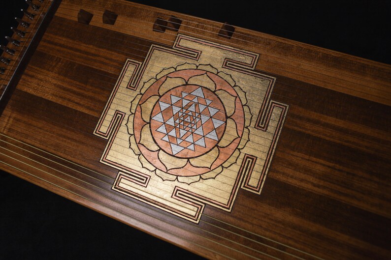 May include: A wooden stringed instrument with an inlaid design. The design features a central geometric pattern in gold, copper, and silver tones, surrounded by a floral-like motif. The instrument's wood grain is visible.