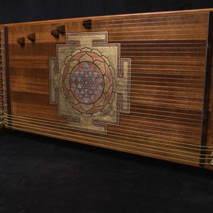 May include: A wooden instrument with a Sri Yantra design. It has many strings, tuning pegs, and a gold, pink, and blue geometric pattern. The wooden frame is a warm brown colour. The instrument is supported by wooden legs.