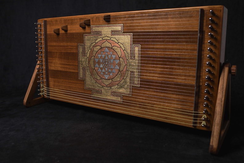 May include: A wooden instrument with a Sri Yantra design. It has many strings, tuning pegs, and a gold, pink, and blue geometric pattern. The wooden frame is a warm brown colour. The instrument is supported by wooden legs.
