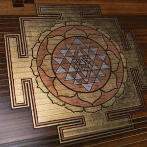 May include: Close-up of a wooden instrument with a decorative design. The design features a central geometric pattern in gold, copper, and silver tones, surrounded by a floral-like shape. The instrument's wood grain is visible.