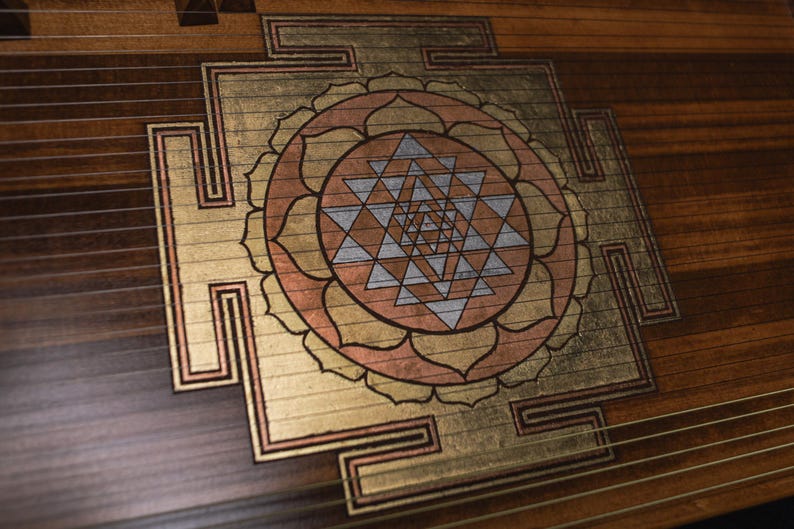 May include: Close-up of a wooden instrument with a decorative design. The design features a central geometric pattern in gold, copper, and silver tones, surrounded by a floral-like shape. The instrument's wood grain is visible.
