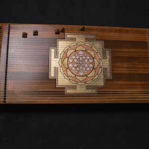 May include: A wooden stringed instrument with a central geometric design in gold, pink, and white. The instrument has a dark brown finish and is set against a black background. It resembles a zither or similar instrument, with many strings.