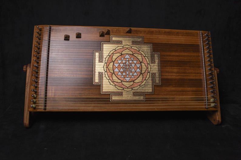 May include: A wooden stringed instrument with a central geometric design in gold, pink, and white. The instrument has a dark brown finish and is set against a black background. It resembles a zither or similar instrument, with many strings.