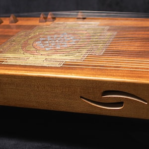 May include: A wooden zither instrument with numerous strings stretched across a rectangular soundboard. The instrument features a decorative inlay and a carved design on the side. The wood has a warm, natural tone.