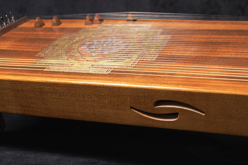 May include: A wooden zither instrument with numerous strings stretched across a rectangular soundboard. The instrument features a decorative inlay and a carved design on the side. The wood has a warm, natural tone.