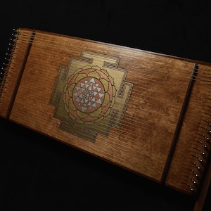 May include: A wooden hammered dulcimer with a decorative geometric design in gold, pink, and silver. The instrument has numerous strings and is set against a dark background. The dulcimer is a stringed instrument.