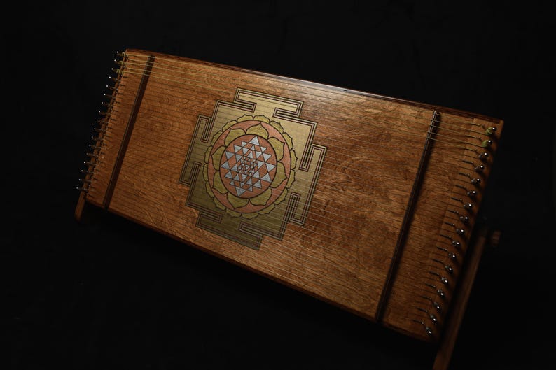 May include: A wooden hammered dulcimer with a decorative geometric design in gold, pink, and silver. The instrument has numerous strings and is set against a dark background. The dulcimer is a stringed instrument.