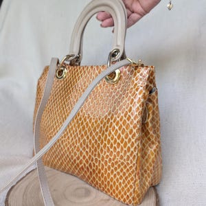 May include: A structured handbag featuring a glossy, amber-coloured snakeskin-patterned exterior. It includes a beige top handle, a matching detachable shoulder strap, and gold-tone metal hardware rings connecting the straps to the bag body.