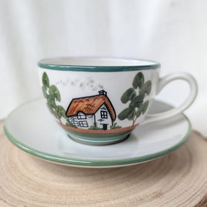 Op de afbeelding: Witte keramische theekop en schotel set met een handgeschilderde huisjesscène. De kop heeft een huisje met een rieten dak, groene bomen en een groene rand. De schotel heeft een bijpassende groene rand. De set staat op een houten oppervlak.