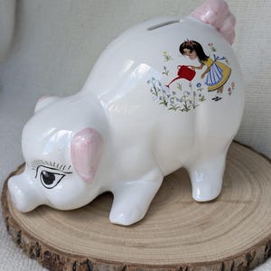 May include: A white ceramic piggy bank with pink painted ears and a pink tail. The side features a colorful illustration of a girl in a yellow dress and blue apron using a red watering can to water a patch of blue and green flowers.