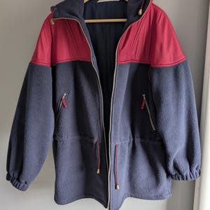 Op de afbeelding: Een marineblauwe fleecejas met een rode capuchon en schouderstuk. De jas heeft een volledige ritssluiting, twee zijzakken en een taille met trekkoord. De jas hangt aan een houten kleerhanger.