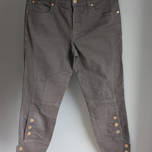 May include: Dark grey pants with gold-colored button details at the ankles. The pants have a tailored fit with a button and zipper closure. The pants are hanging on a black hanger.