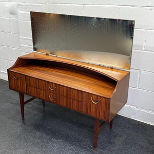 May include: A vintage wooden dressing table with a rectangular mirror. The table has a warm brown finish, three drawers, and curved brass handles. It stands on four tapered legs.