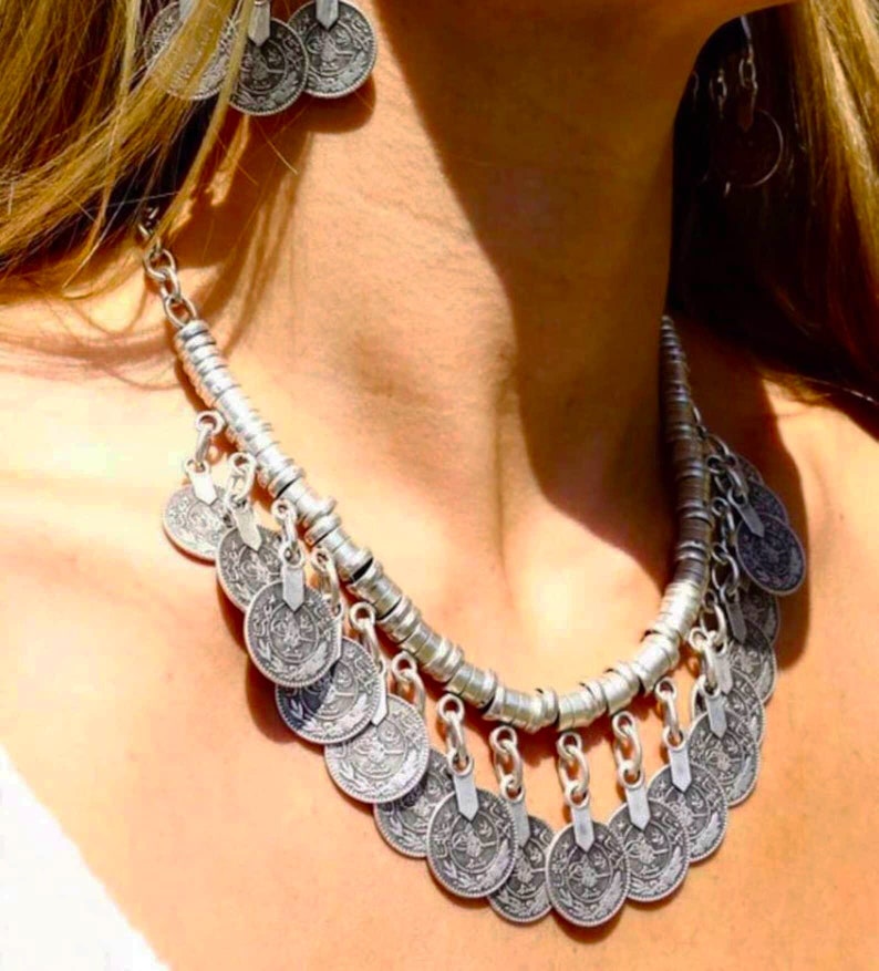 May include: A silver necklace with a chain and multiple coin-shaped pendants. The necklace is worn by a person with long blonde hair.