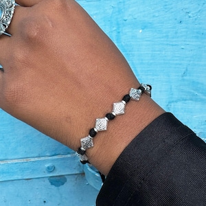 May include: A black cord bracelet with silver charm beads. Each bead features a decorative, geometric design.