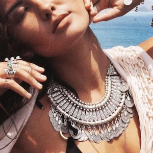 Könnte beinhalten: Eine silberne Statement-Kette mit einem großen, verzierten Anhänger und mehreren münzenförmigen Charms. Die Kette wird über einem weißen Oberteil getragen und von einer Person mit dunklen Haaren getragen.