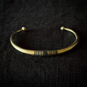 Pode incluir: Uma pulseira de metal em tom dourado com um design curvo e aberto, apresentando terminais arredondados. O centro da banda exibe uma secção escura e texturizada com múltiplas linhas verticais finas gravadas, contrastando com o acabamento dourado liso.