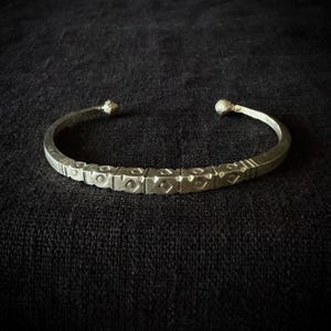 Pode incluir: Uma pulseira de metal em tom prateado com uma série de segmentos retangulares na parte frontal, cada um gravado com um formato de diamante central. A pulseira tem um design aberto em forma de C com extremidades arredondadas e texturizadas.