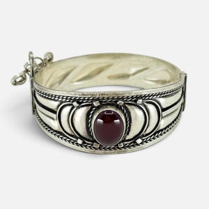 Pode incluir: Uma pulseira de metal prateado com uma pedra preciosa oval de cor vermelho-escura no centro. A banda é decorada com padrões semicirculares em relevo e bordas de corda torcida. Um pequeno mecanismo de fecho metálico está fixado num lado.