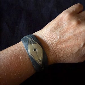 Pode incluir: Uma pulseira de punho cinza-escura texturizada com uma placa metálica central em forma de diamante com acabamento polido. A placa apresenta um padrão de linhas gravadas e uma pequena pedra escura circular no centro, fixada com costuras metálicas visíveis.