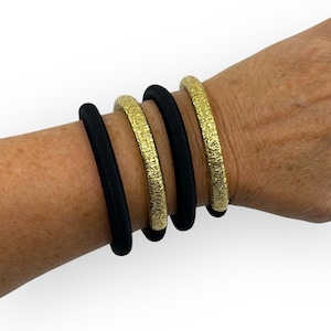 Peut inclure: Un poignet orné de quatre bracelets. Deux sont noirs et lisses, tandis que les deux autres sont dorés avec un aspect texturé et martelé. Les bracelets sont portés ensemble, créant un look superposé. Le fond est blanc uni.
