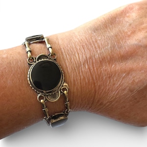 Peut inclure: Un bracelet composé d'une grande pierre noire ronde et polie, sertie dans une monture en métal aux tons laiton texturé. La pierre centrale est flanquée de pierres noires rectangulaires plus petites, reliées par des maillons métalliques.