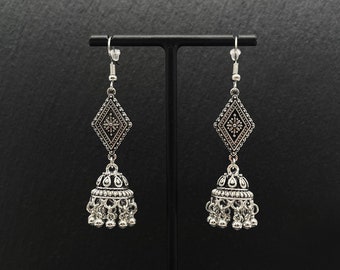 May include: A pair of silver dangle earrings with a geometric design and a bell-shaped charm. The earrings are hanging from a black stand.