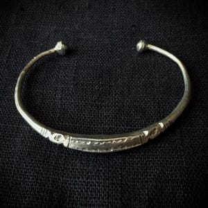Könnte beinhalten: Ein silberfarbener Metall-Armreif mit offenem Design und abgerundeten Enden. Der mittlere Teil des Bandes weist ein texturiertes, geätztes Muster mit geometrischen Markierungen und eine leicht abgeflachte, längliche rechteckige Form auf.