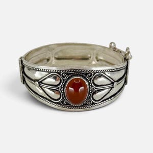 Pode incluir: Uma pulseira de metal prateado com uma pedra central oval de cor castanho-avermelhada. A banda é decorada com formas em relevo semelhantes a corações e detalhes metálicos em estilo de corda torcida. Inclui um mecanismo de fecho articulado.